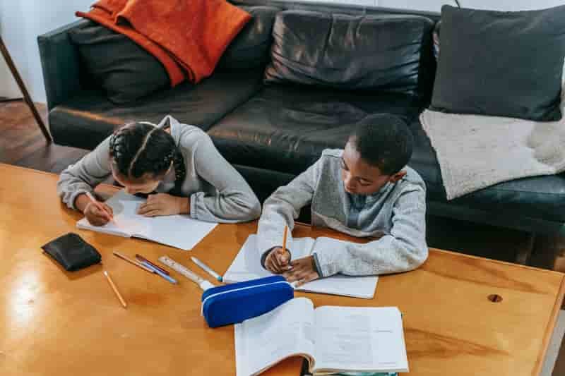 Two primary-school children doing homework together at a wooden table, with notebooks, pencils and a textbook spread between them.