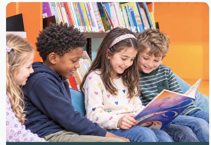 Four children smiling and reading a picture book together in a colorful school library.