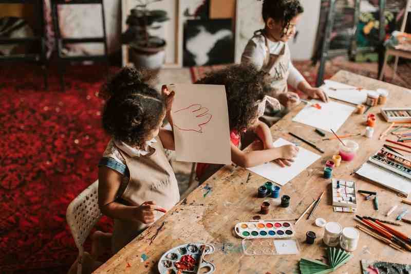 Three children painting at a big wooden studio table, one holding up a drawing of her own hand.
