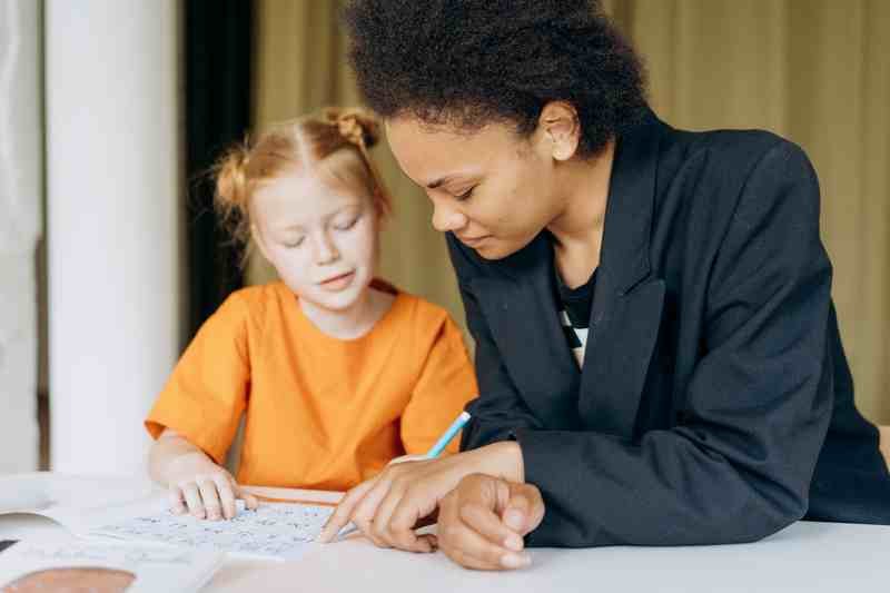 A tutor leaning in beside a young girl, both looking at the same letters worksheet.