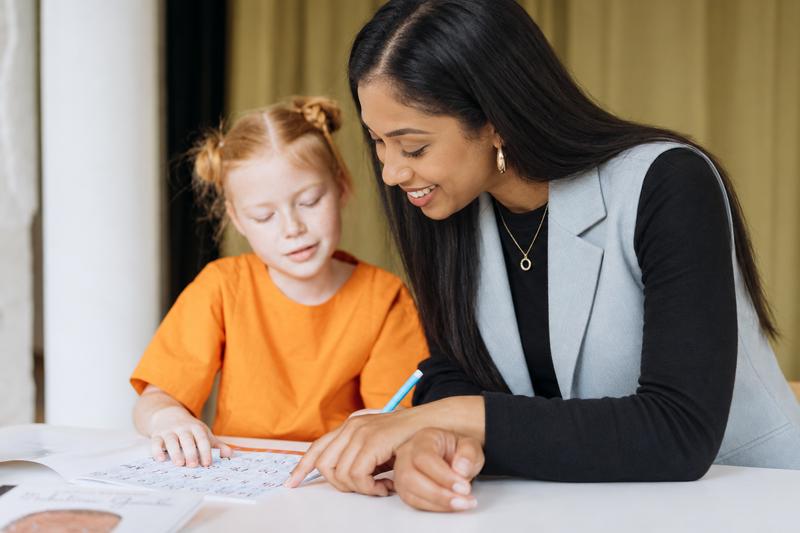 A tutor leaning in beside a young girl, both looking at the same letters worksheet.